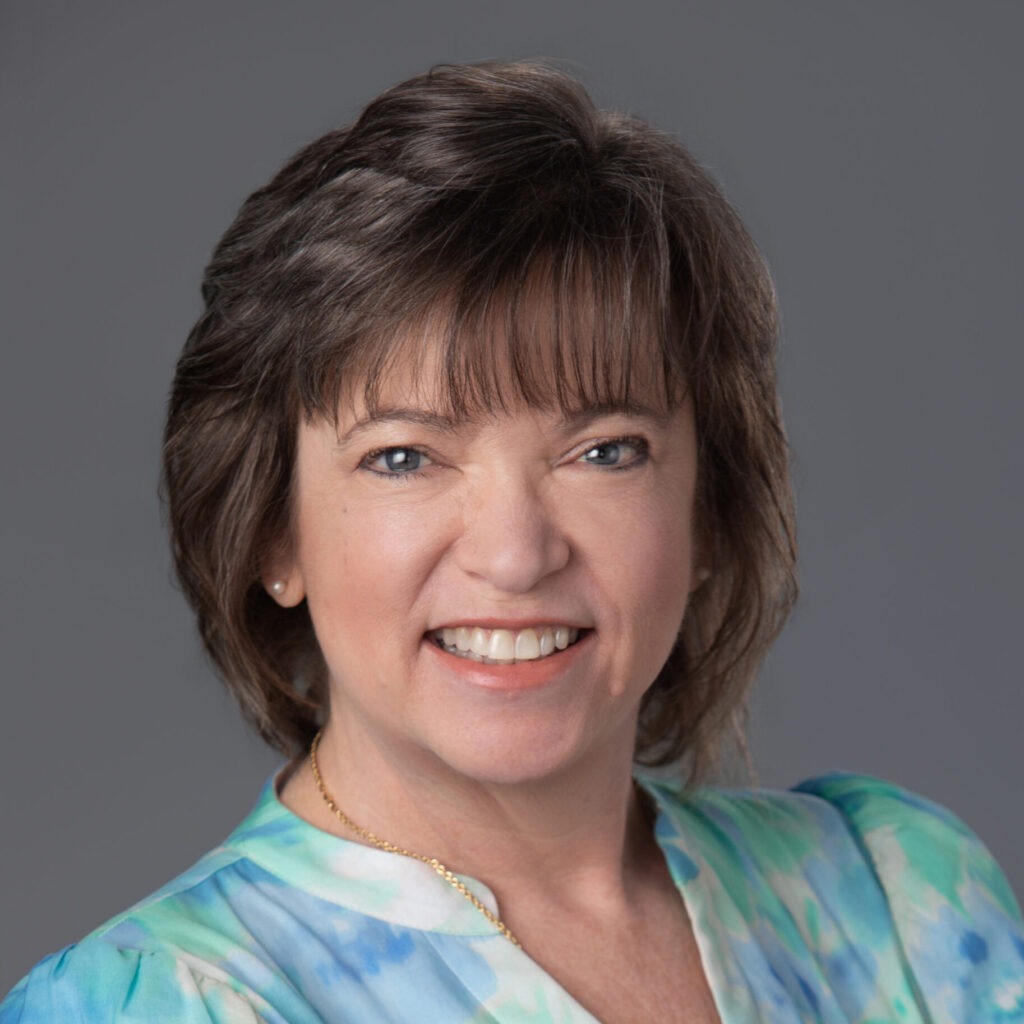 Portrait of a smiling woman with short brown hair and blue eyes, wearing a blue-green tie-dye blouse against a gray background.
