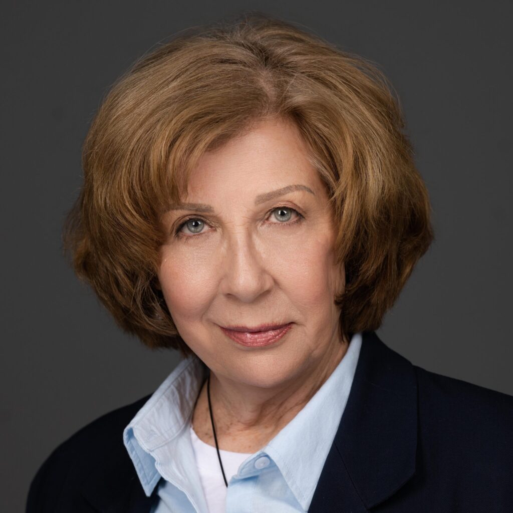 Close-up portrait of a smiling older woman with short light brown hair, wearing a navy blazer and light blue shirt against a gray backdrop.